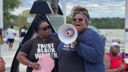 Angela Aina and Monica Simpson stand in front of the Liberty Bell, Aina holding a megaphone during a speech at a rally.