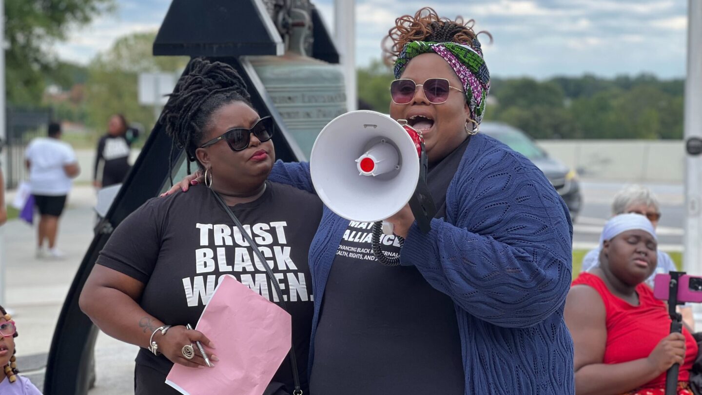 Angela Aina and Monica Simpson stand in front of the Liberty Bell, Aina holding a megaphone during a speech at a rally.