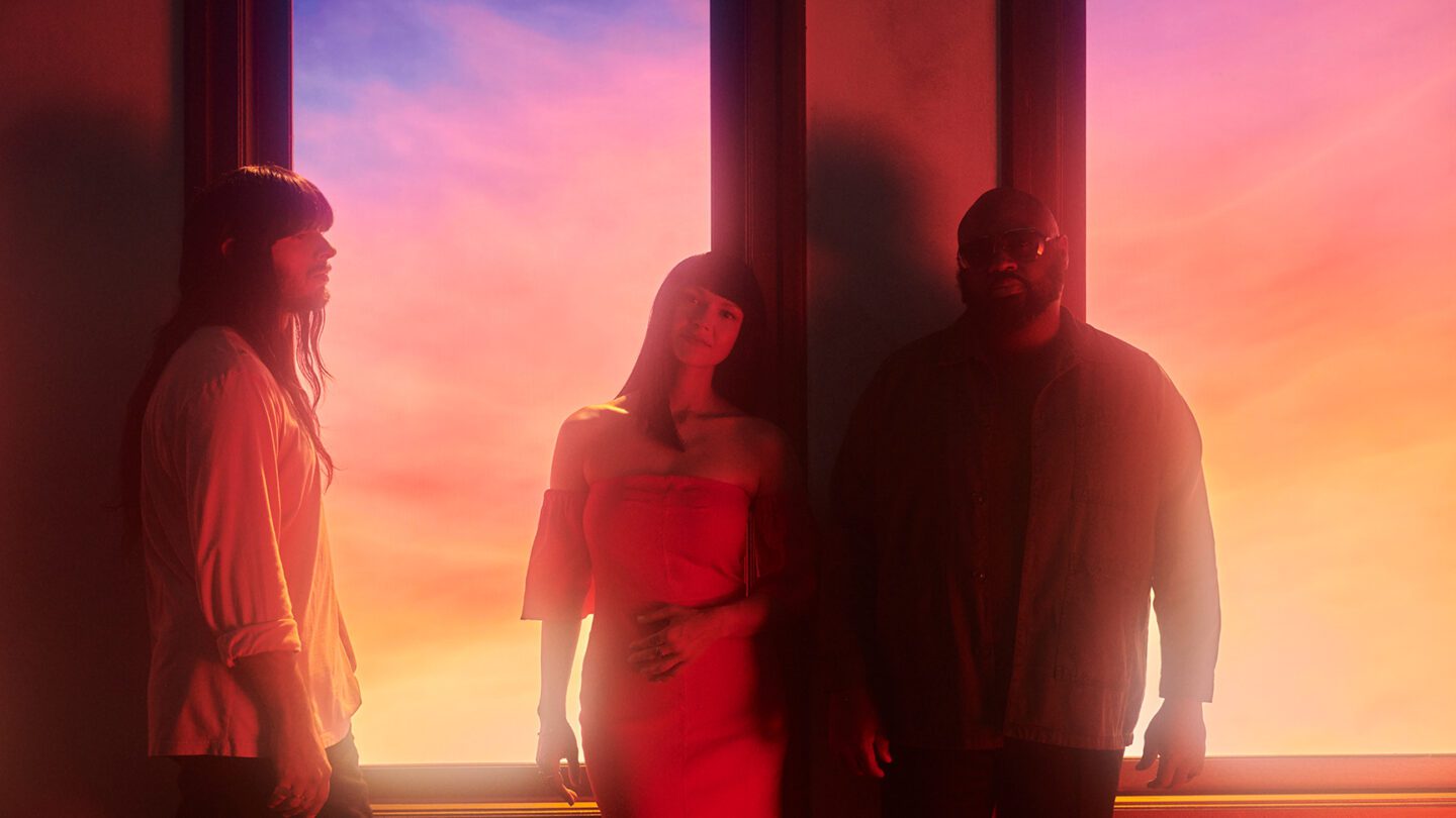 A photo of Khruangbin standing in front of a window with a colorful sunset in the background.