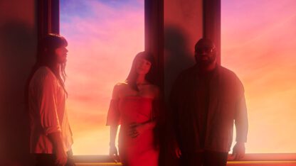 A photo of Khruangbin standing in front of a window with a colorful sunset in the background.