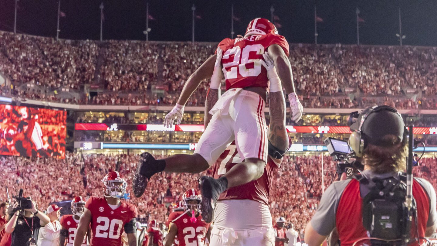 A football player raises another football player in the air in the middle of a crowded stadium.