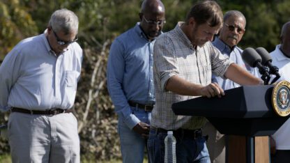 USDA Secretary Tom Vilsack stands on the left and lowers his head as pecan farmer Buck Paulk leads a prayer during a press event for Helene recovery in Ray City, Georgia.