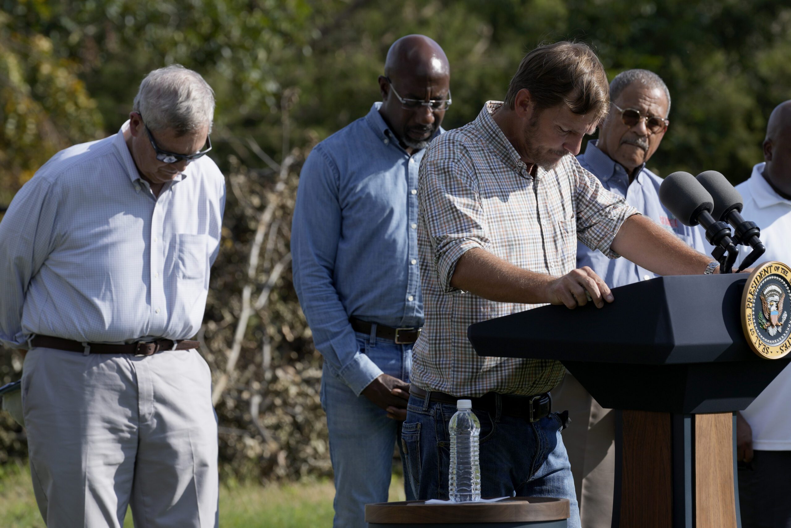 USDA Secretary Tom Vilsack stands on the left and lowers his head as pecan farmer Buck Paulk leads a prayer during a press event for Helene recovery in Ray City, Georgia.