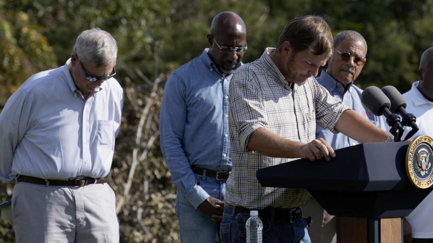 USDA Secretary Tom Vilsack stands on the left and lowers his head as pecan farmer Buck Paulk leads a prayer during a press event for Helene recovery in Ray City, Georgia.