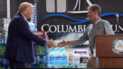 Donald Trump, left, and Brian Kemp, right shake hands at press event, standing next to cases of water and boxes of diapers and other supplies.