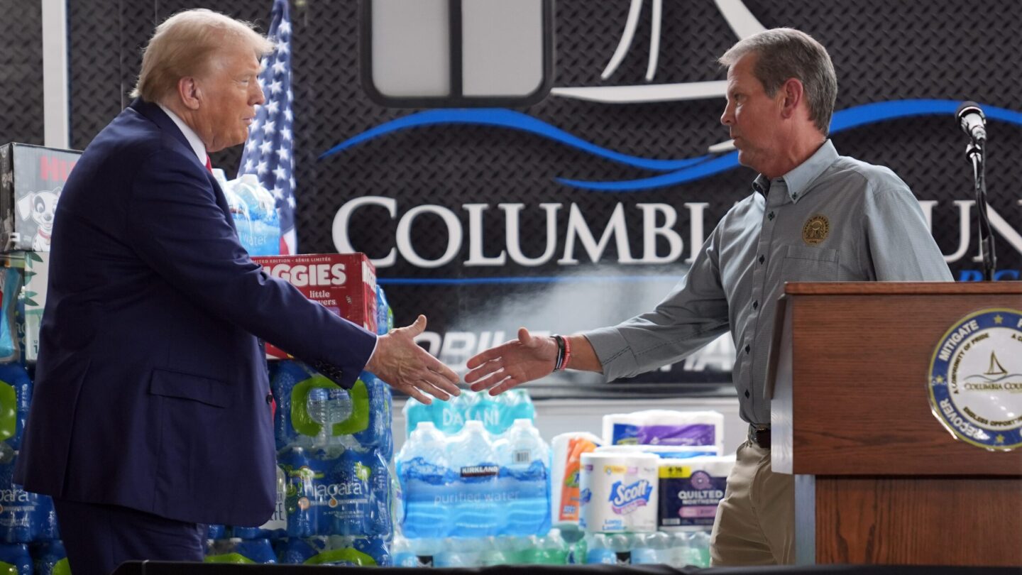 Donald Trump, left, and Brian Kemp, right shake hands at press event, standing next to cases of water and boxes of diapers and other supplies.