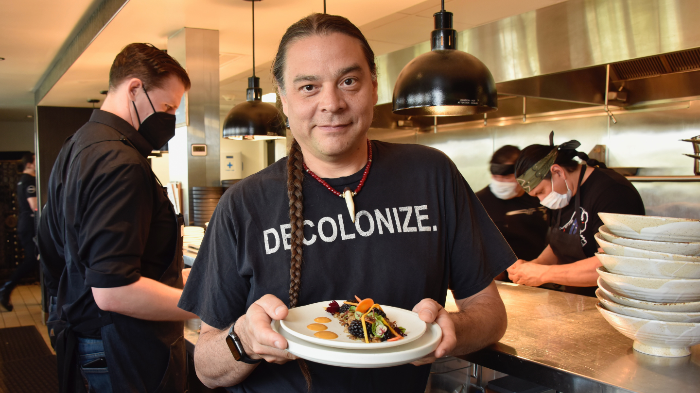 Chef Sean Sherman poses with a dish in a kitchen. He wears a shirt that says Decolonize.