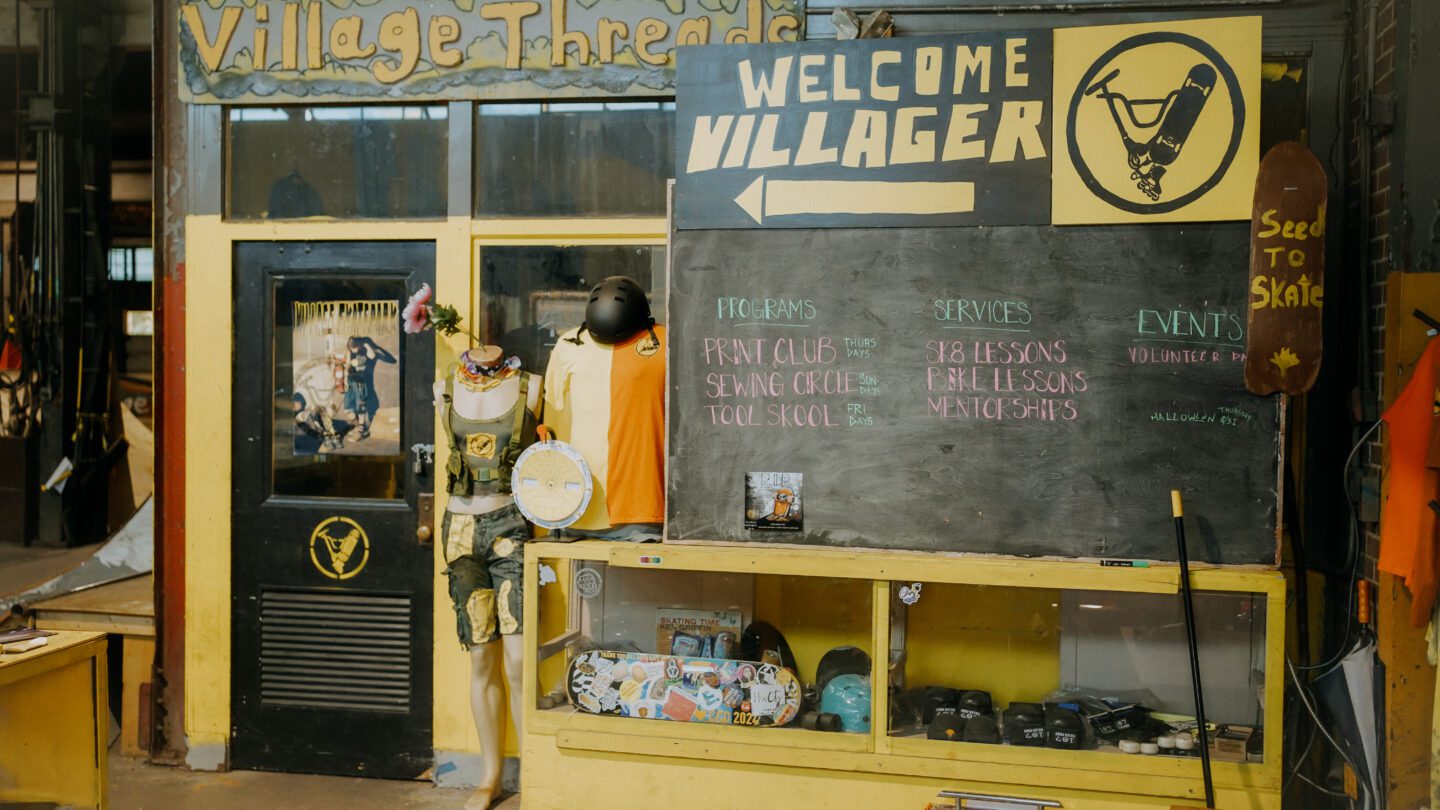 A small indoor storefront that says Village Threads, with a bulletin board in front of it that shows a variety of programming.