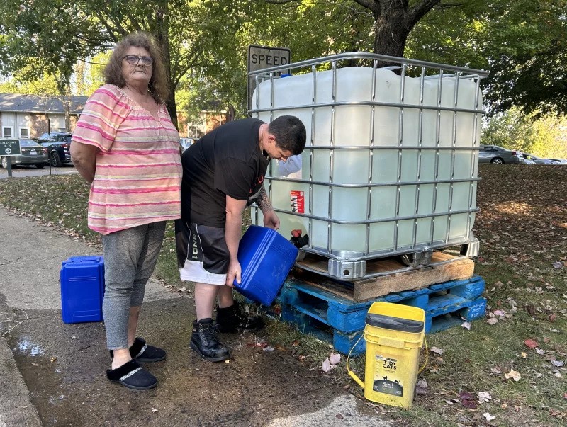 Teresa Thomas stands to the left as her son bends down to fill up buckets with water from a large tank.