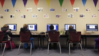 Students sitting at a row of desks and using desktop computers.