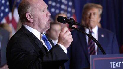 Tom Homan faces right, speaking, as President-elect Donald Trump looks on in the background.