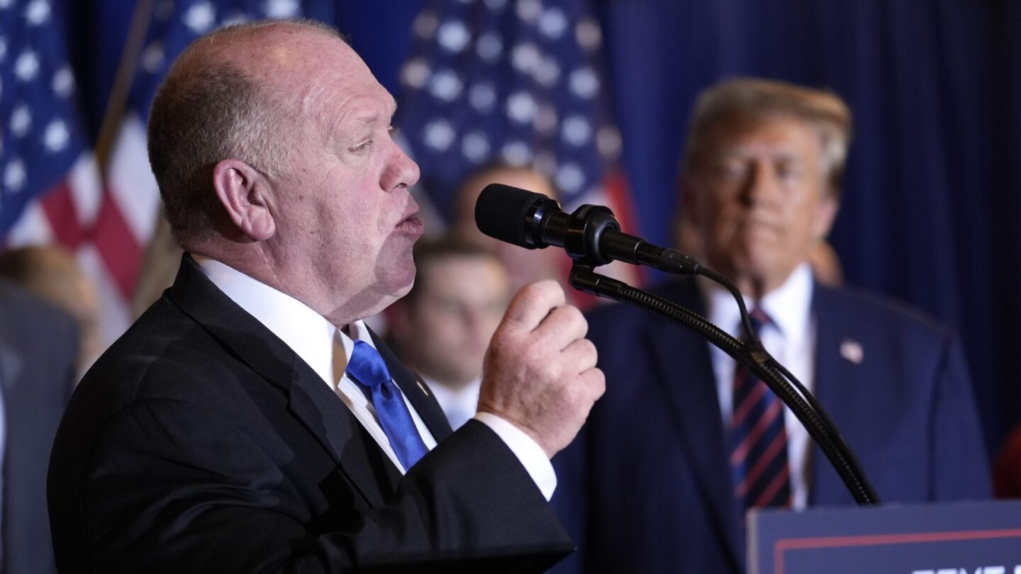 Tom Homan faces right, speaking, as President-elect Donald Trump looks on in the background.