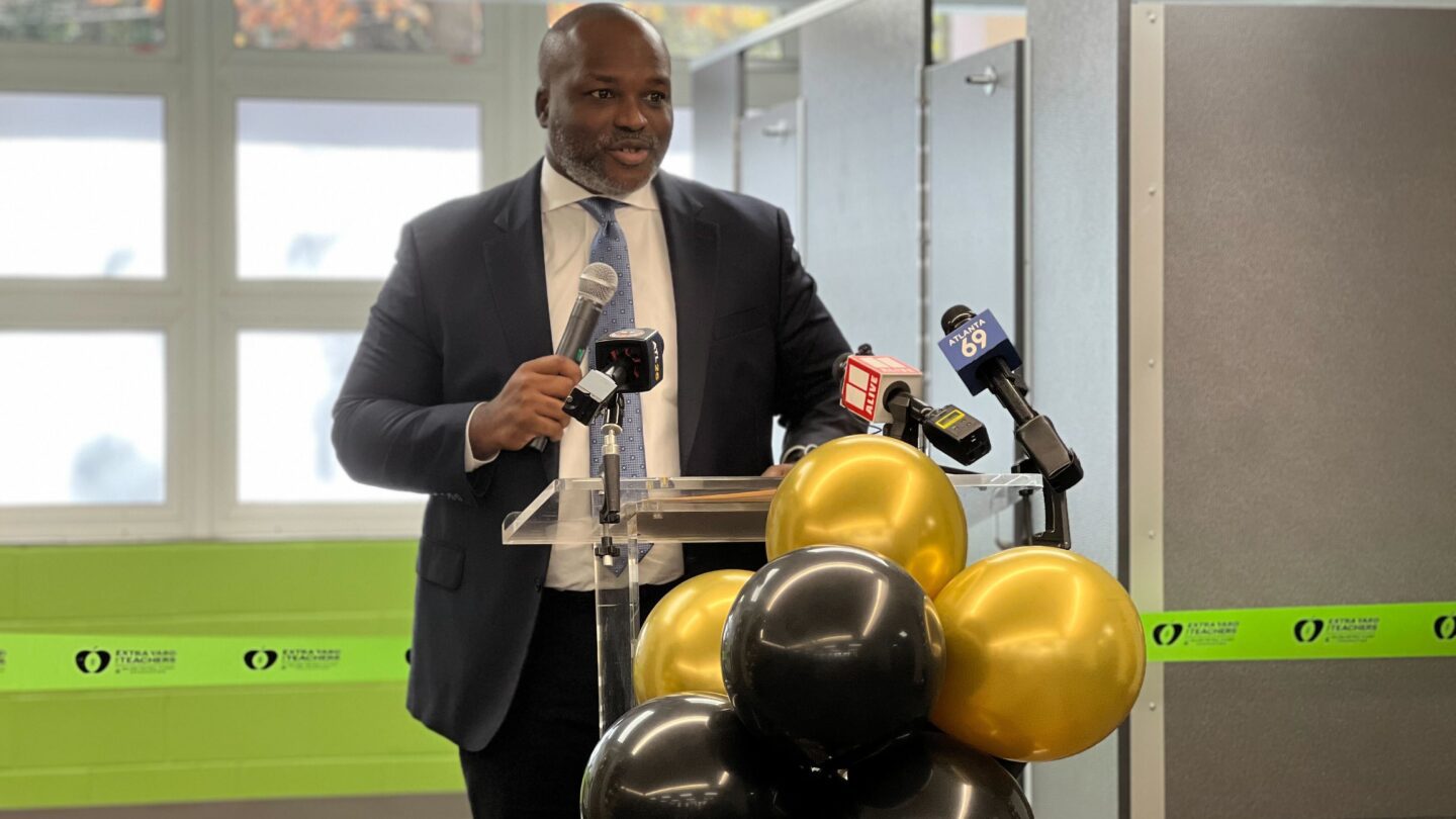 Atlanta Public Schools Superintendent Bryan Johnson speaks at a clear podium with black and yellow balloons, holding a microphone, standing in front of a green ribbon.