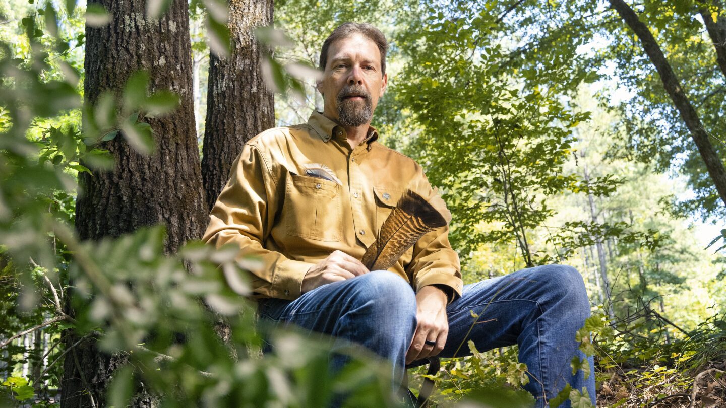 Mike Chamberlain in a yellow long sleeve shirt and jeans sits in the forest, surrounded by greenery, holding a wild turkey feather.