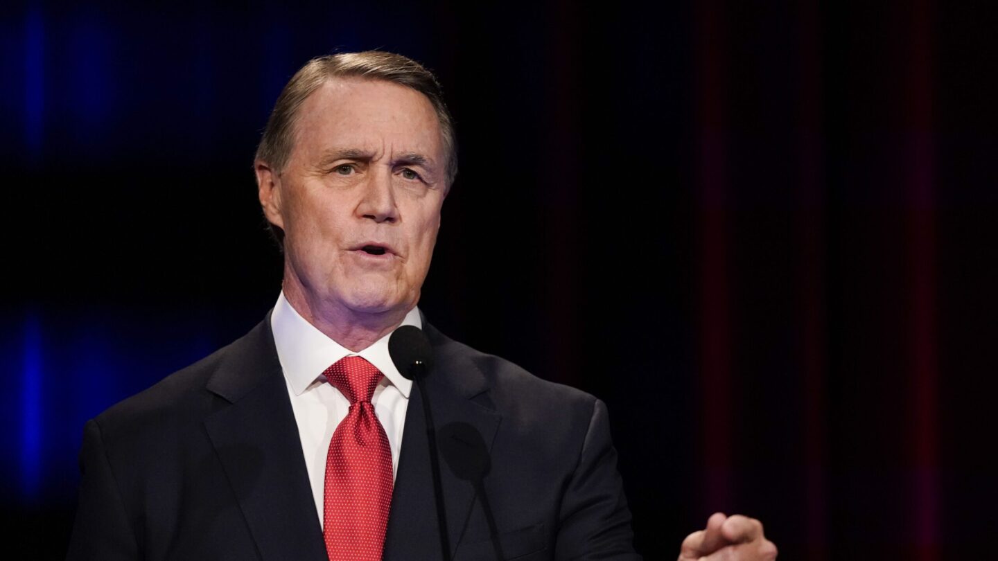 David Perdue wearing a red tie speaking at a podium.