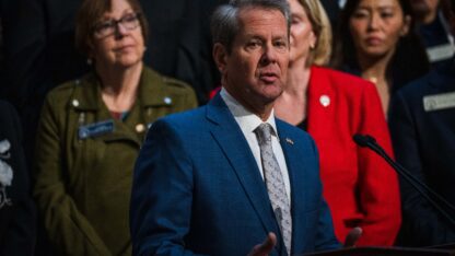 Kemp stands in front of a group of legislators at the Georgia State Capitol, speaking at a podium.