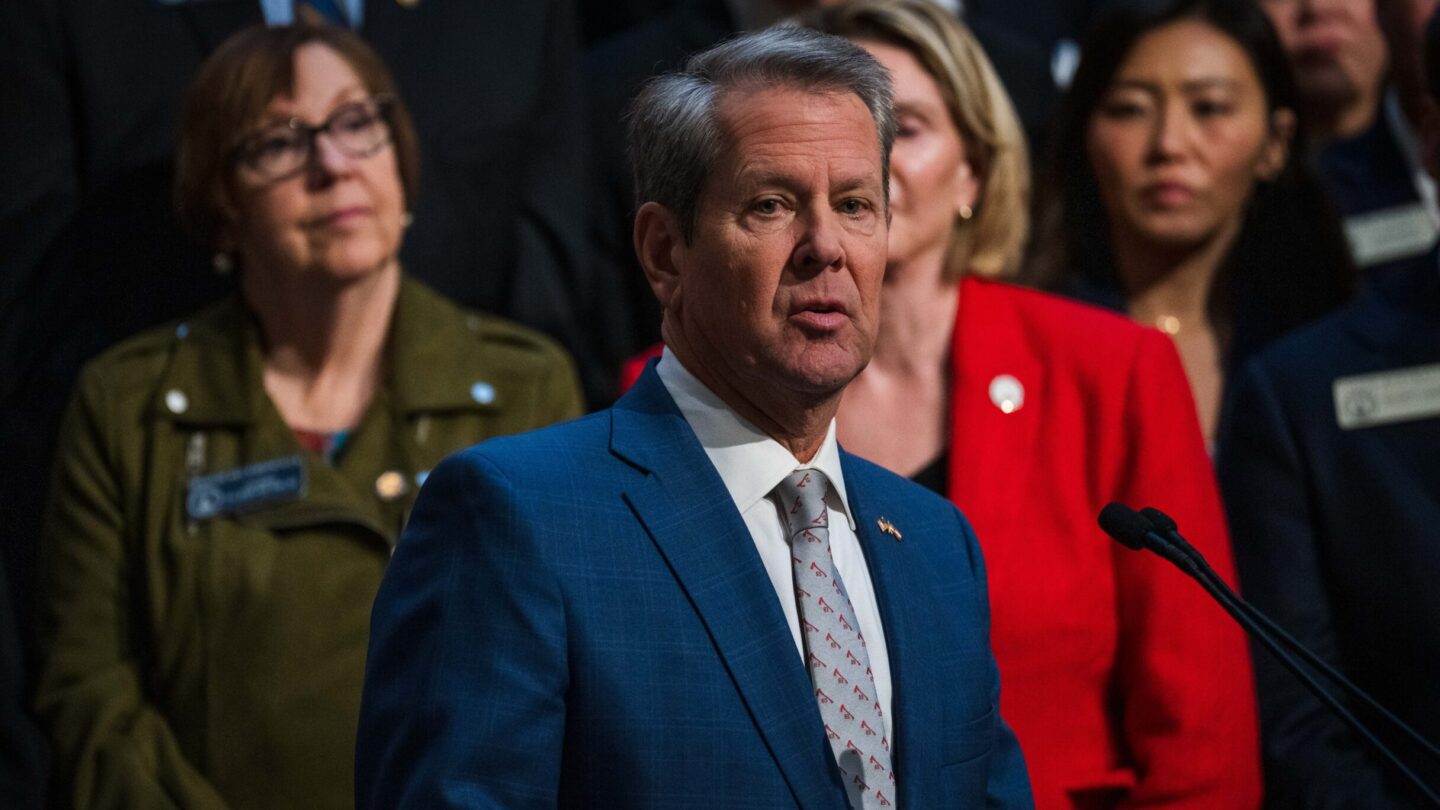 Kemp stands in front of a group of legislators at the Georgia State Capitol, speaking at a podium.