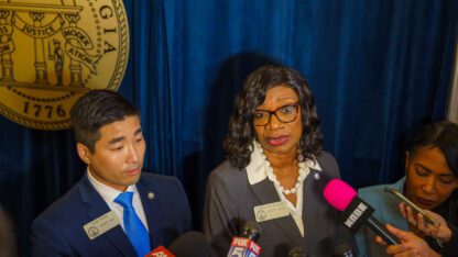 Democratic leaders Sam Park, Carolyn Hugley, and Tanya Miller stand in front of a blue curtain in front of a crowd of microphones.