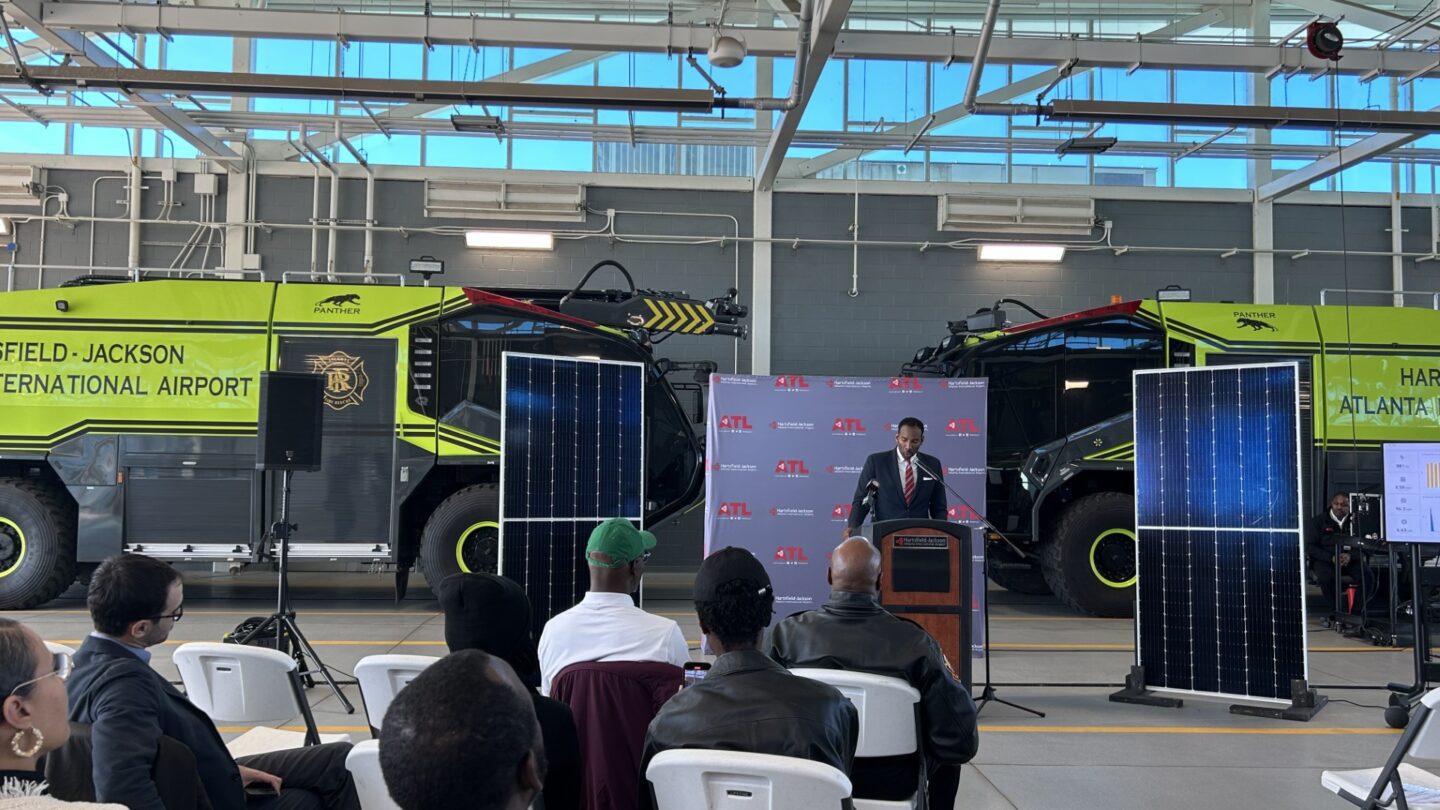 Mayor Andre Dickens stands behind a podium in front of two large green trucks. In front of him are a few rows of chairs in an arch, facing away from the photographer.