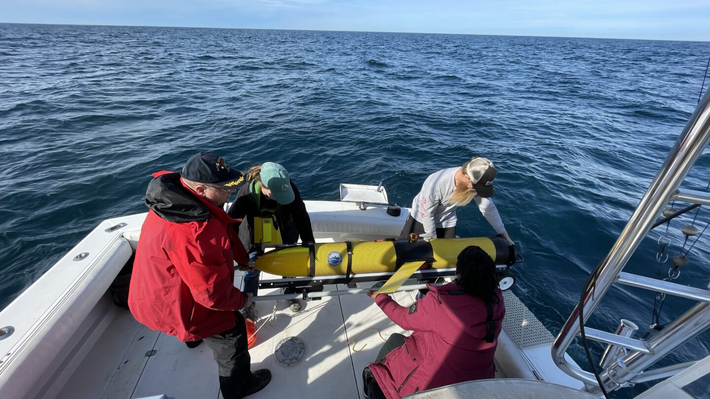 A group of people on a boat holds a bright yellow, torpedo-shaped glider